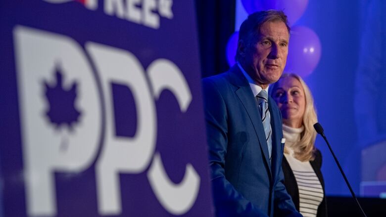 A man stands at a podium delivering a speech. In the foreground is a People's Party of Canada sign.