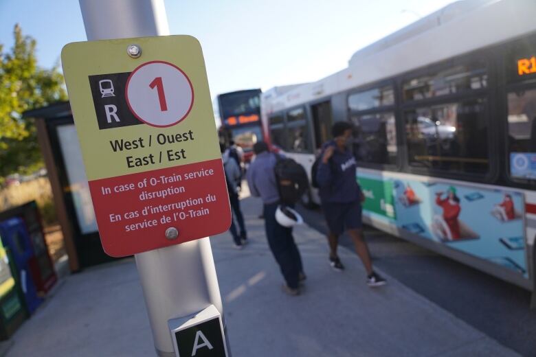 A yellow and red sign describes replacement bus service. In the background people walk by a bus.