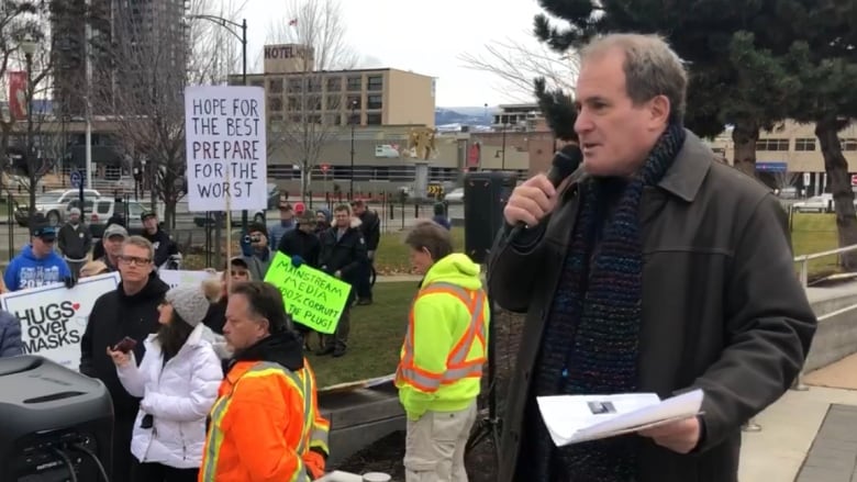 A photo of a middle-aged man holding a microphone and speaking to a rally of several dozen people at a public square in Kelowna, B.C. 