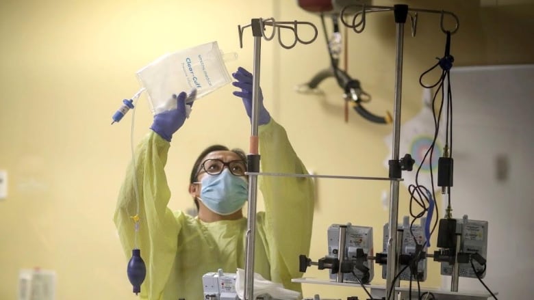 A health-care worker wearing a blue medical mask, yellow gown and blue surgical gloves, replaces an IV bag of fluid on a metal pole overhead.