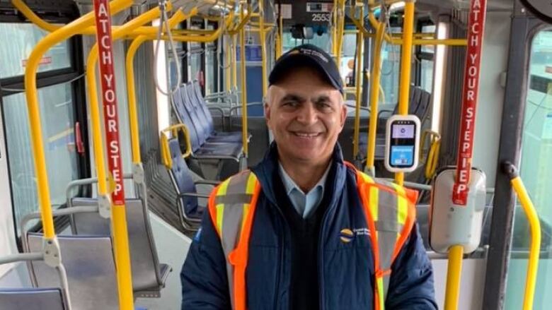 A man in a safety vest and ball cap stands in the middle of a city bus, smiling at the camera 