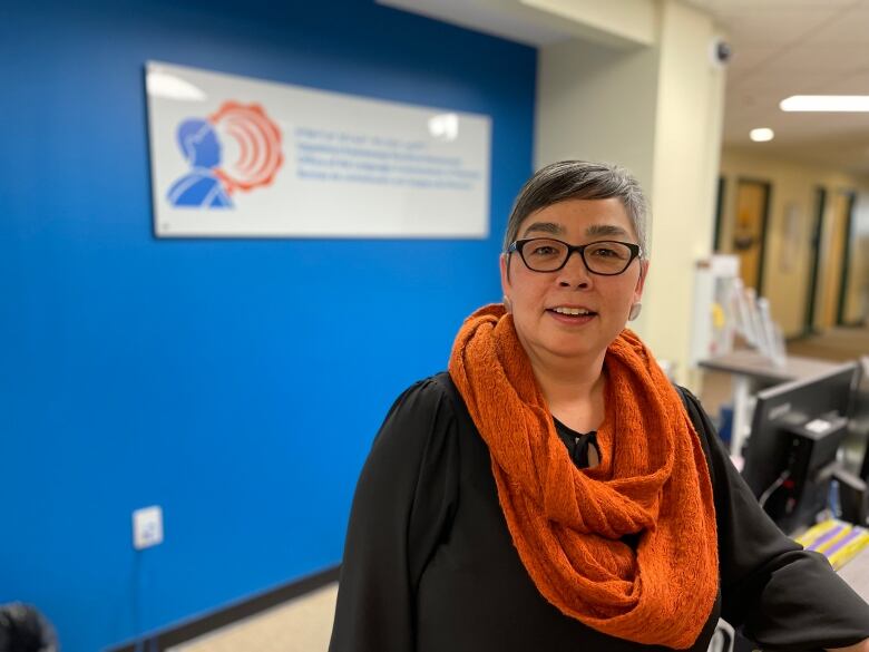 A woman with short grey hair wearing glasses, a black shirt and an orange scarf