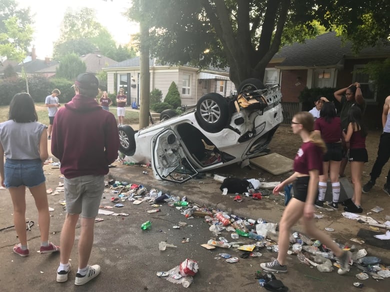 Young people stand around a car that has been flipped upside down on a sidewalk.