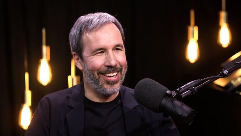 A man with grey hair and a beard sits in front of a microphone in a room lit with bare light bulbs. 