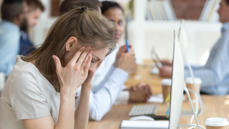 A visibly upset woman holds her head while sitting in front of a computer in an office, surrounded by people.