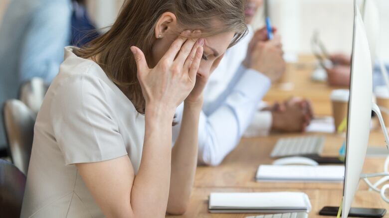 A visibly upset woman holds her head while sitting in front of a computer in an office, surrounded by people.