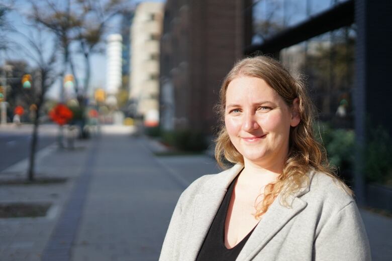 A woman wearing a grey suit jacket stands outside on the sidewalk in an urban setting.