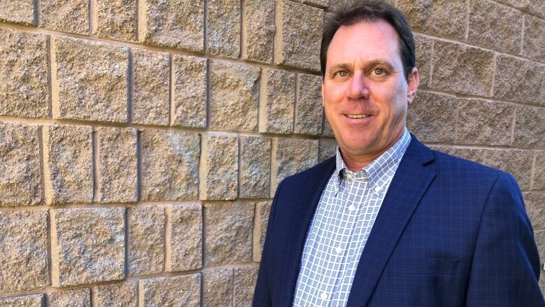 A man in a blue blazer and a blue checked shirt poses for a photo in front of a brick wall.