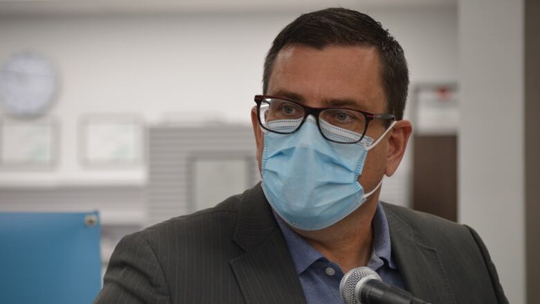 A man in glasses and a suit jacket wears a blue face mask as he sits in front of a microphone.