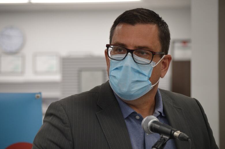 A man in glasses and a suit jacket wears a blue face mask as he sits in front of a microphone.