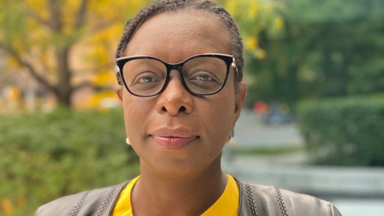 A middle aged black woman in business attire stands in a park, looking at the camera. It's daytime. She's visible from the waist up.