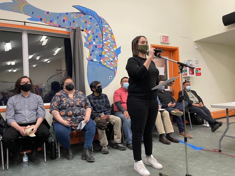 A woman with short hair and wearing a white mask stands in the foreground with a microphone, while a row of people sit in chairs behind her. 