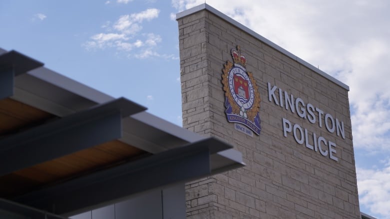 A sign for Kingston Police, along with their logo, can be seen on a grey brick wall. The sky in the background is blue with some clouds.