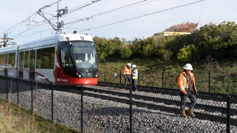 Workers in bright orange walk along some rail tracks, with a white and red light-rail train car behind them. 