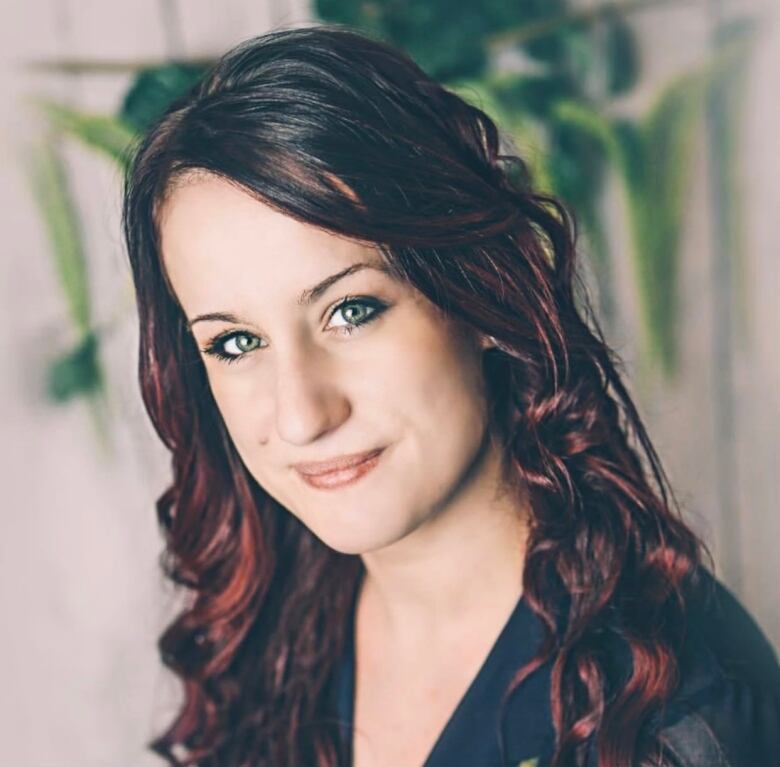 A three quarters head shot of a woman with flowing brown hair with reddish highlights and a plant in the background.