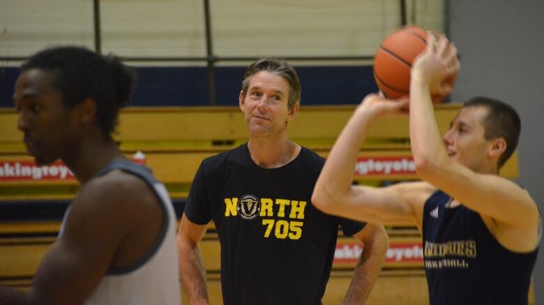 In a gym, a basketball player takes a shot in practice while his coach watches from the sideline. A third player is visible on the left side of the picture.