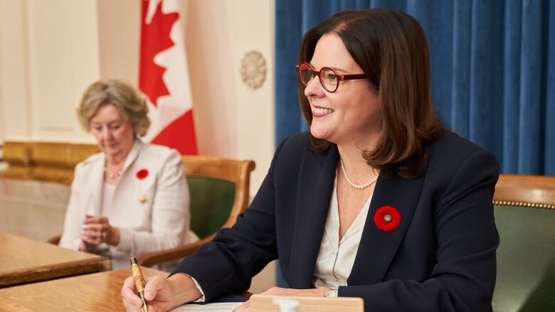 A woman in a dark suit and white blouse smiles while seated as she prepares to sign a document. 