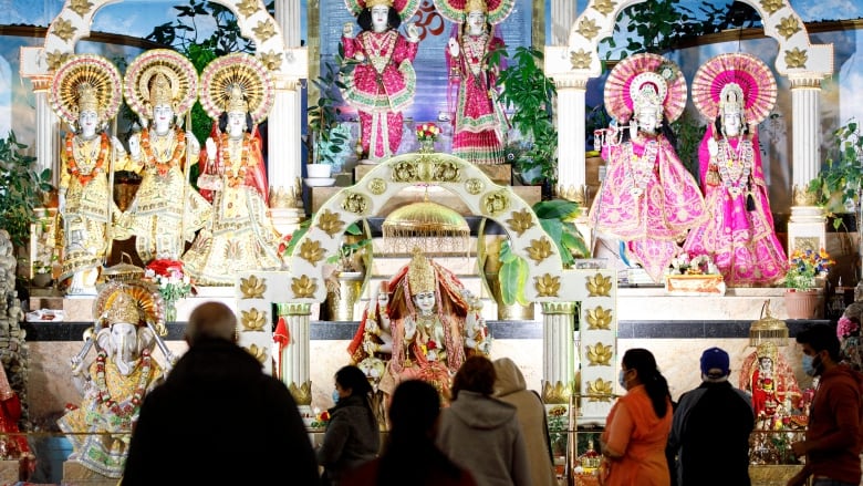 People standing at a place of worship with filled with statues wearing colourful clothing and grand decorations