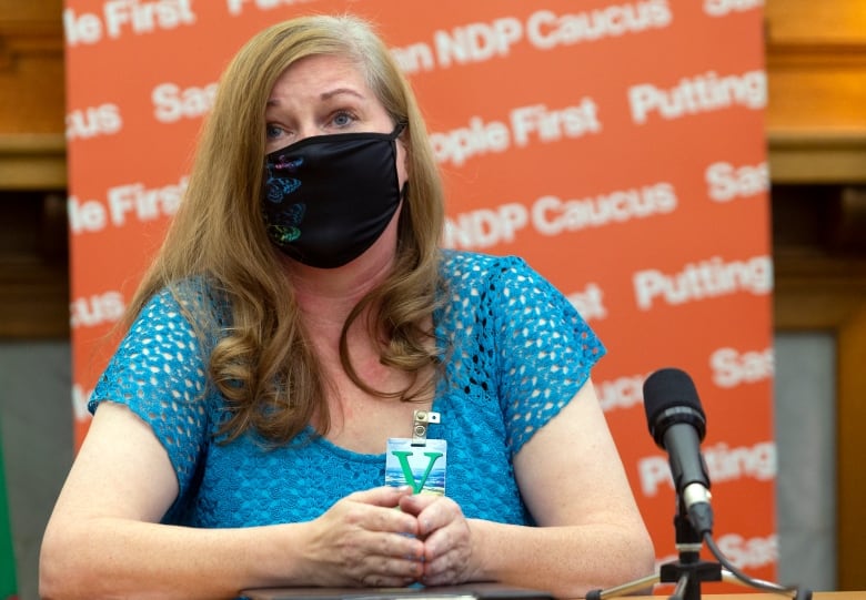 A blonde woman wearing a facemask speaks to a mic while sitting in front of an orange banner.