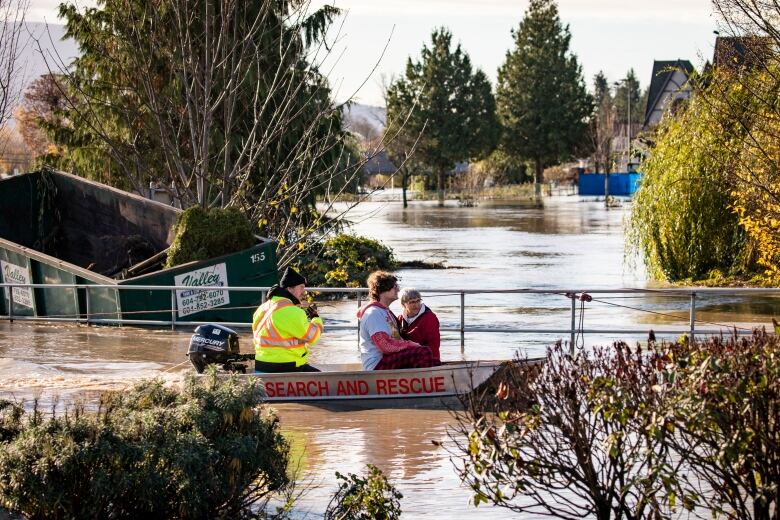 Three people travel on a boat marked 'Search and Rescue' amid heavy floods in a residential area.