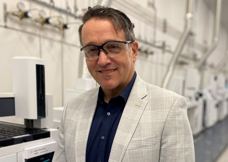 Man in a dark blue shirt and cream checked jacket wears safety glasses while standing in a factory. 