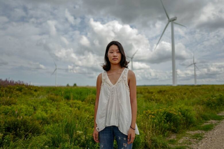 A young woman stands on a trail in a grassy field. Large wind turbines can be seen behind her in the distance, among a cloudy blue sky.