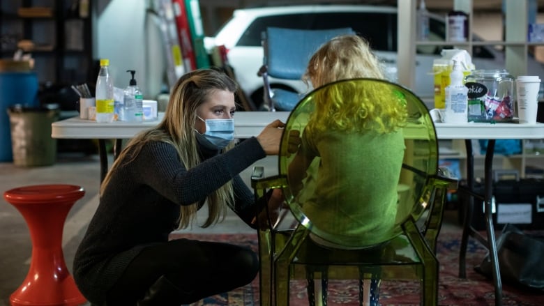 A nurse in a face mask bends down to give a child, seated in a chair, a vaccination.