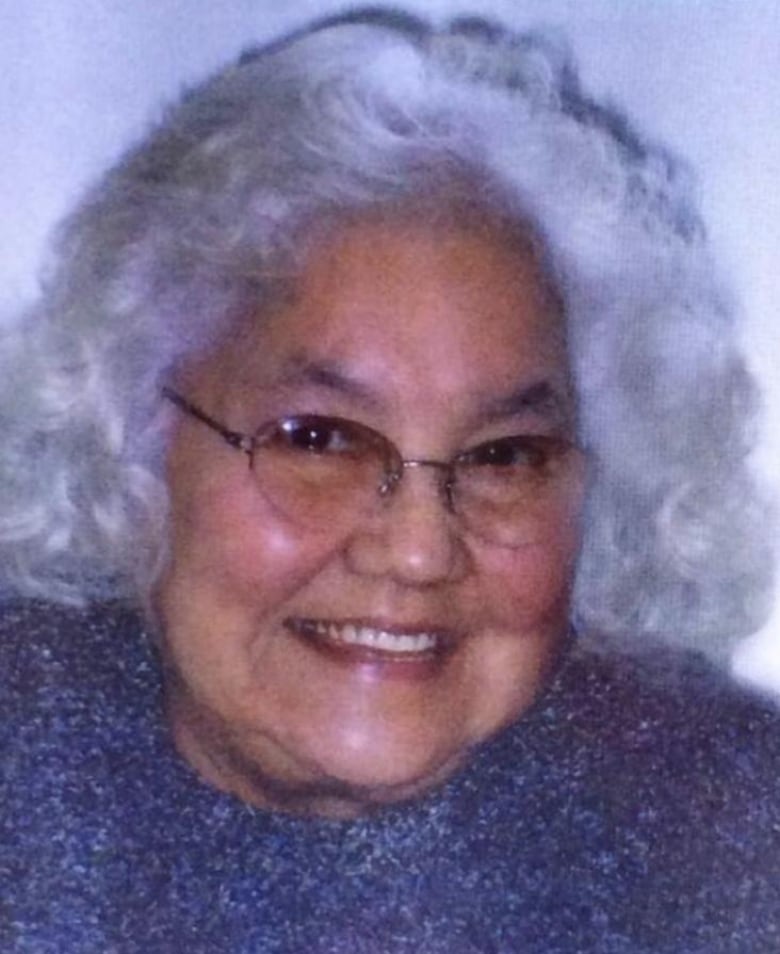 A woman with white hair and glasses, and a blue sweater, poses for a photo.