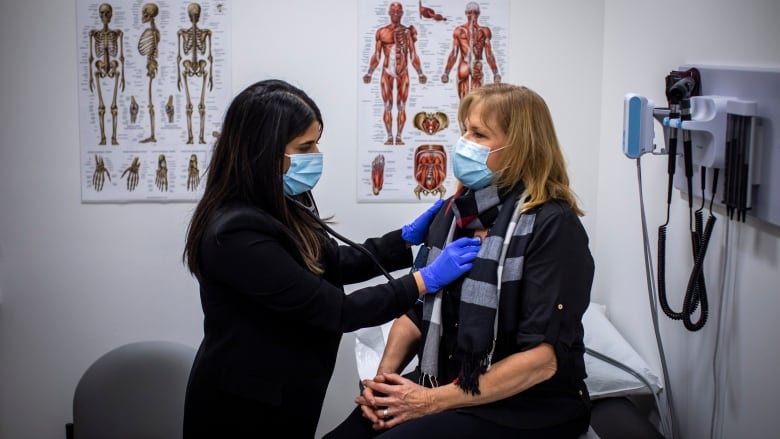 A nurse practitioner listens to a patient's heart with a stethoscope. Both are wearing masks.