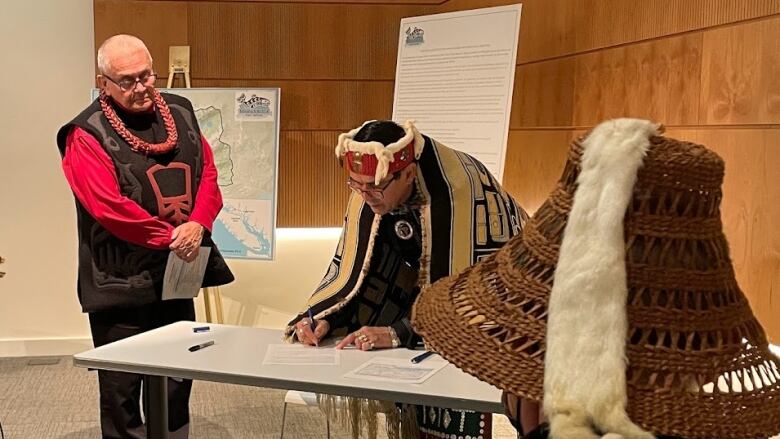 A man in traditional Indigenous regalia signs an agreement while others watch in a meeting room.