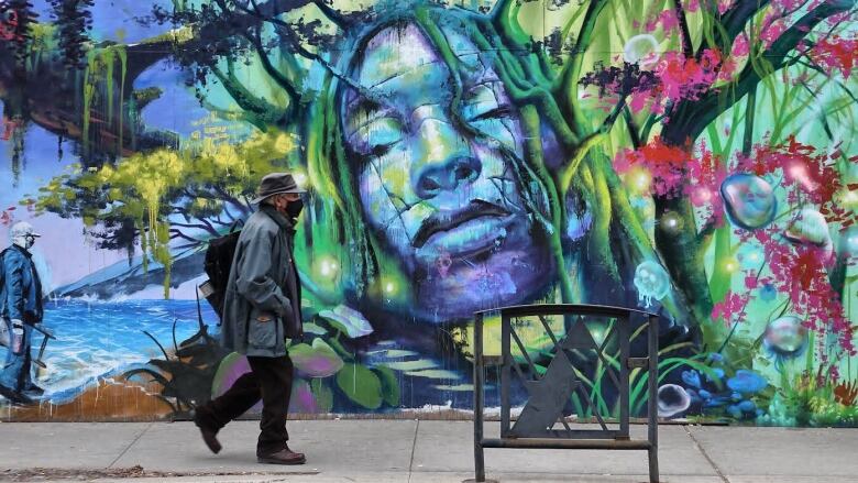 A man walks past a city street mural with a nature scene and a blue face on it. It's late autumn.