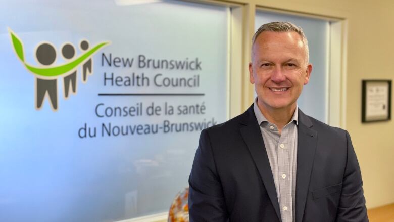 A man with short gray hair, wearing a dark suit and light gray dress shirt smiles at the camera for an upper body portrait standing in front of an opaque window with the words New Brunswick Health Council on it and a logo featuring three humanoid figures and a bright green swish connecting them.