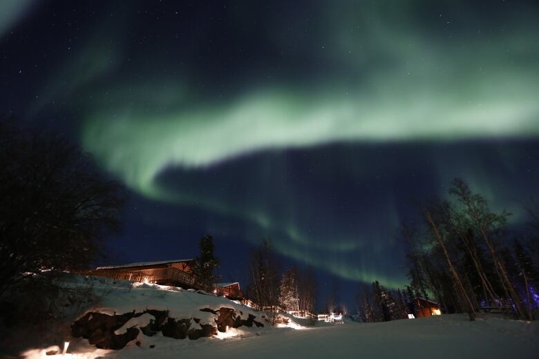 Colourful northern lights dance across the sky as a lodge sits in the background on a snowy, rocky terrain. 