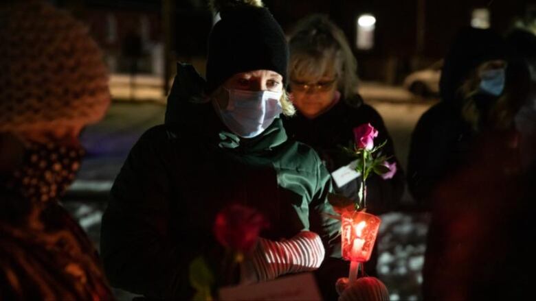 Someone in a mask and winter clothes holds a candle at a nighttime vigil.