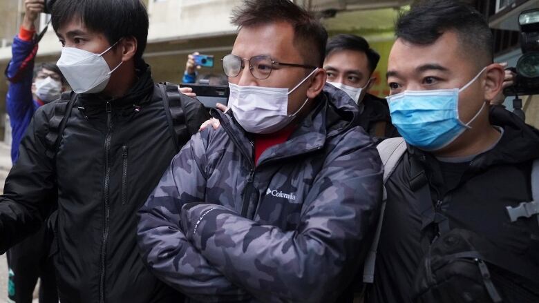 A man wearing glasses and a surgical mask is flanked by two police officers.