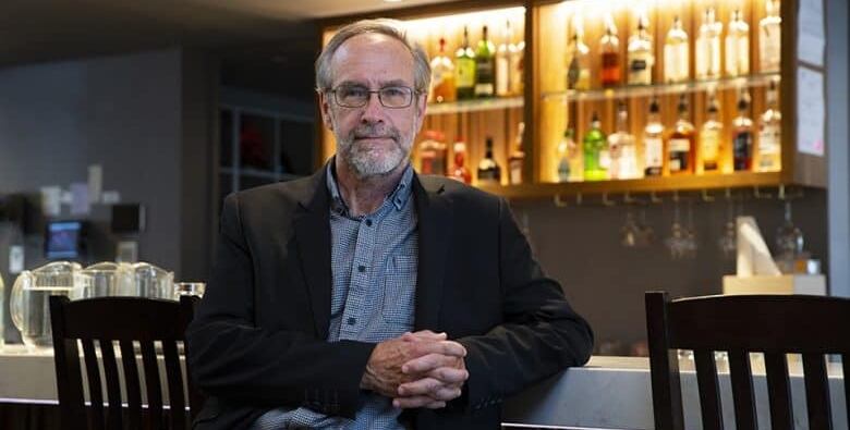 An older man with a beard and glasses sits in front of a lit liquor shelf and looks at the camera.