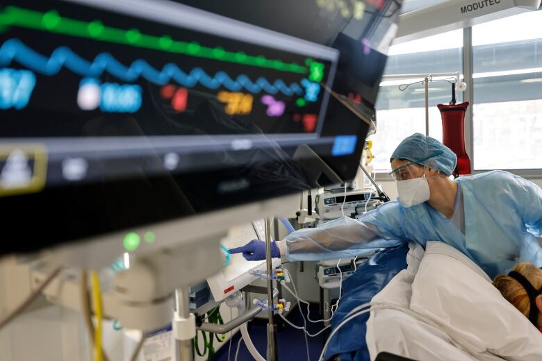 A nurse reaches toward a piece of medical equipment beyond the top of a patient's bed, while a monitor is seen in the foreground.