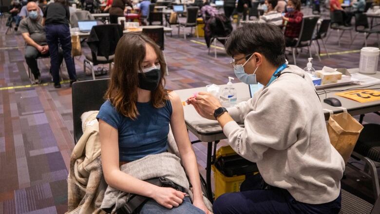 A girl receives a vaccine in a convention hall. Behind her, dozens more people are seen getting vaccinated.