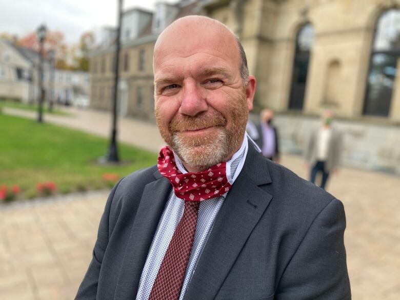 A man in a blue suit and red tie stands outside smiling at the camera.