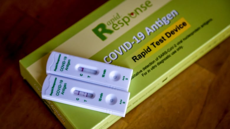 Two COVID rapid tests sit on top of a green box, marked 'Rapid Response, COVID-19 Antigen Rapid Test Device.' One has two lines -- meaning it is positive. The other has one line, meaning it is negative.