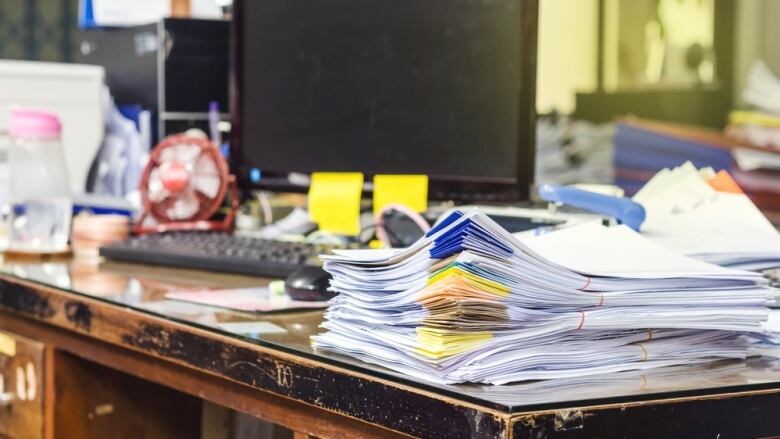 A stack of folders and documents sit on a weather wooden desk in front of a computer.