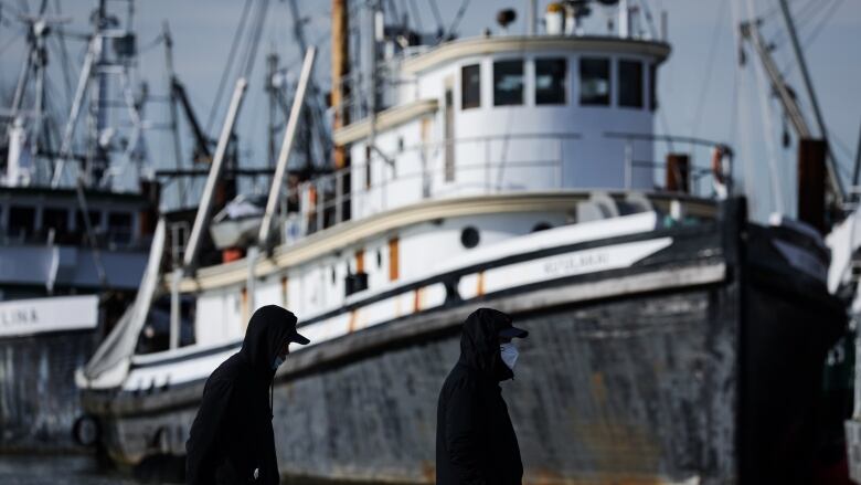 People wearing face masks to curb the spread of COVID-19 walk past a commercial fishing vessel docked at Steveston Village, in Richmond, B.C., Saturday, Jan. 22, 2022.