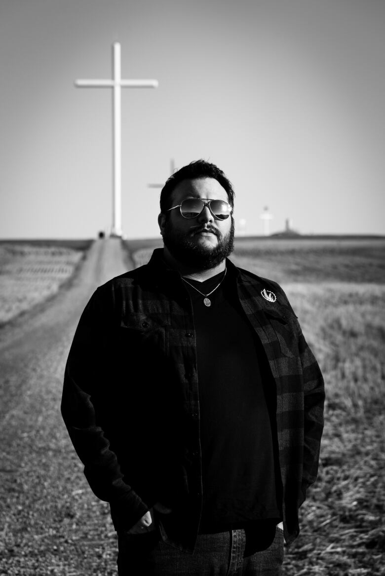 A black & white photo of a Mexican man standing on a dirt road while wearing shades