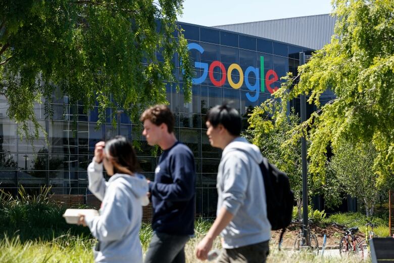 Three people walk by a large building with the Google logo on it.