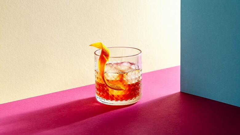 glass with whiskey and ice cubes on a bright pink surface with hard shadows. the walls behind it are blue and beige. 