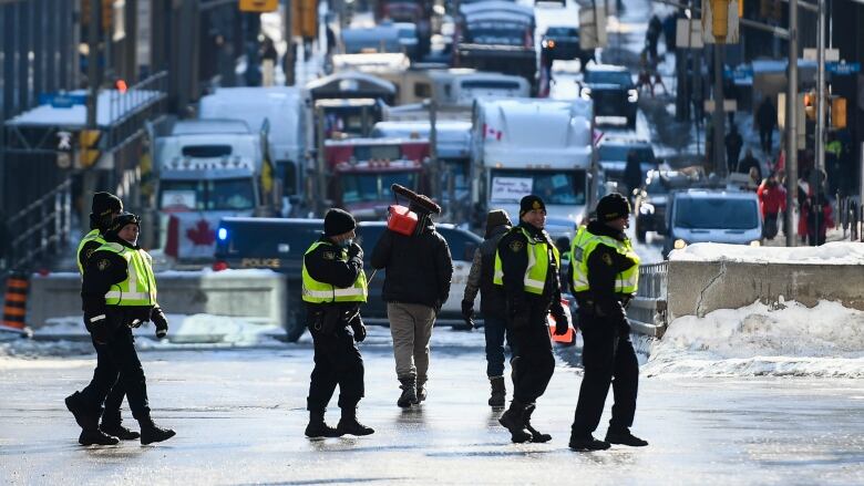 A small group of police officers in yellow vests walk past a long line of trucks parked on a city street. Someone carries a fuel can past the officers.