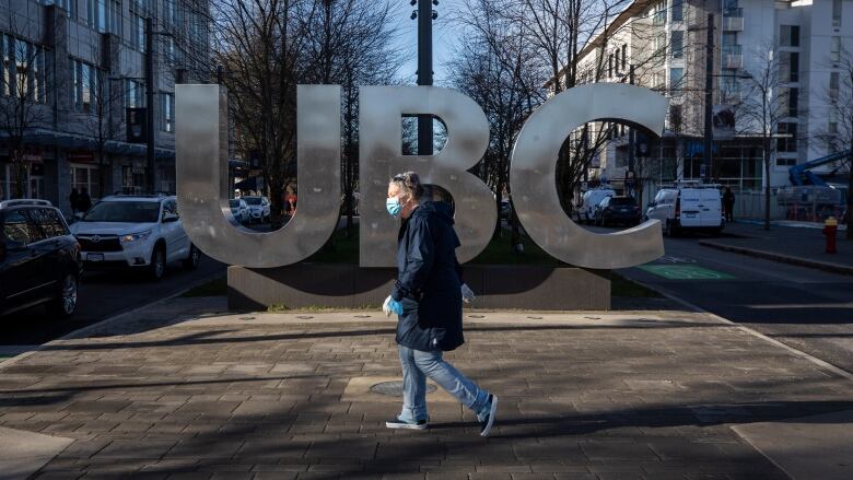 A pedestrian wears a facemask while crossing by a sign that reads 'UBC'.