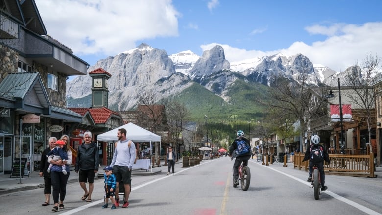 Tourists are seen walking in the middle of the street when cars were not allowed in the resort town of Canmore, with shops on either side of the road and mountains in the background.