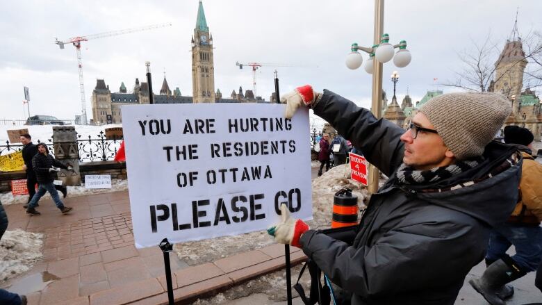 A man attaches a sign to a Parliament Hill fence saying 'You are hurting the residents of Ottawa, please go.'