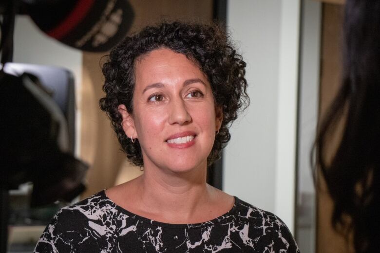 A woman with dark, curly hair listens to questions from a reporter.